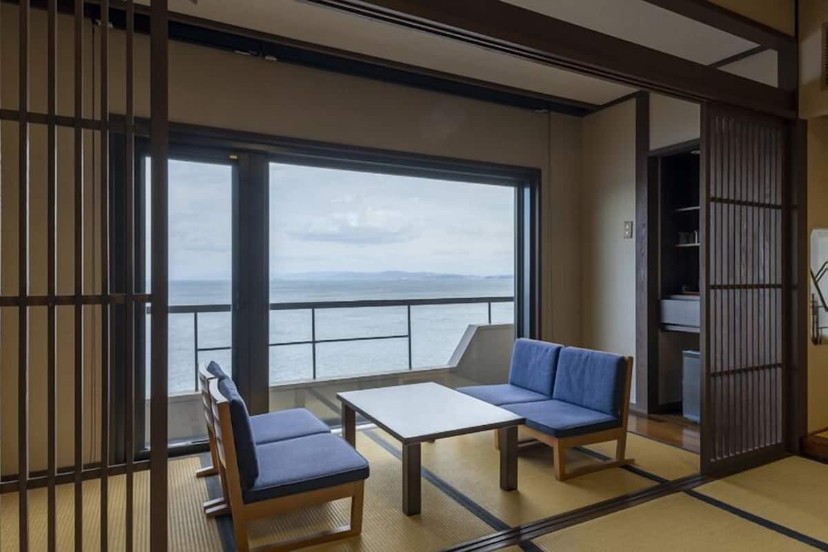 Ein Zimmer mit Tatami-Boden, einem niedrigen Tisch und zwei blauen Sitzgelegenheiten, sowie ein großes Fenster mit Meerblick.