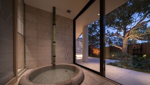 Un moderno espacio de spa con un jacuzzi de piedra, una fuente de agua de bambú y vistas a un jardín arbolado a través de amplias puertas de cristal.