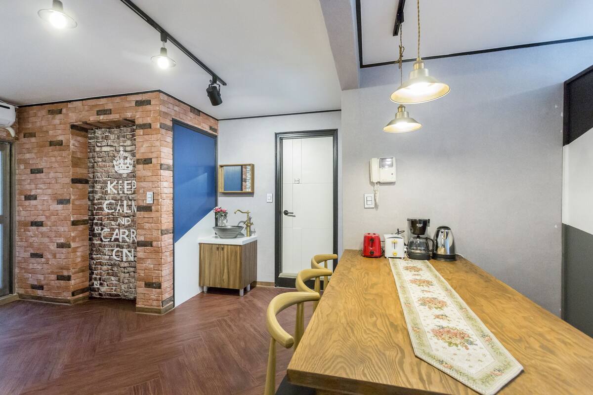 A modern kitchen with a wooden table, hanging lights, and a brick accent wall.