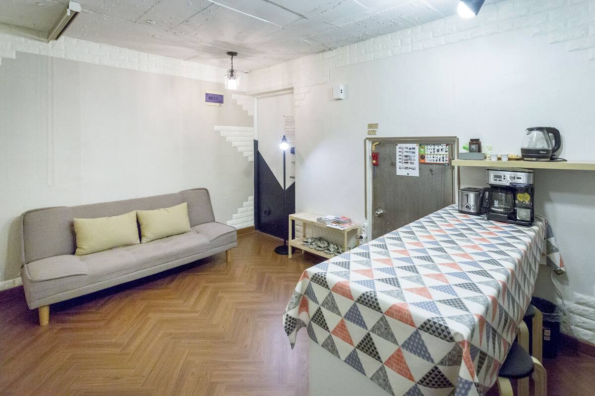 A room with a sofa, a bed with a patterned bedspread, a coffee machine, and a shelf with various items.