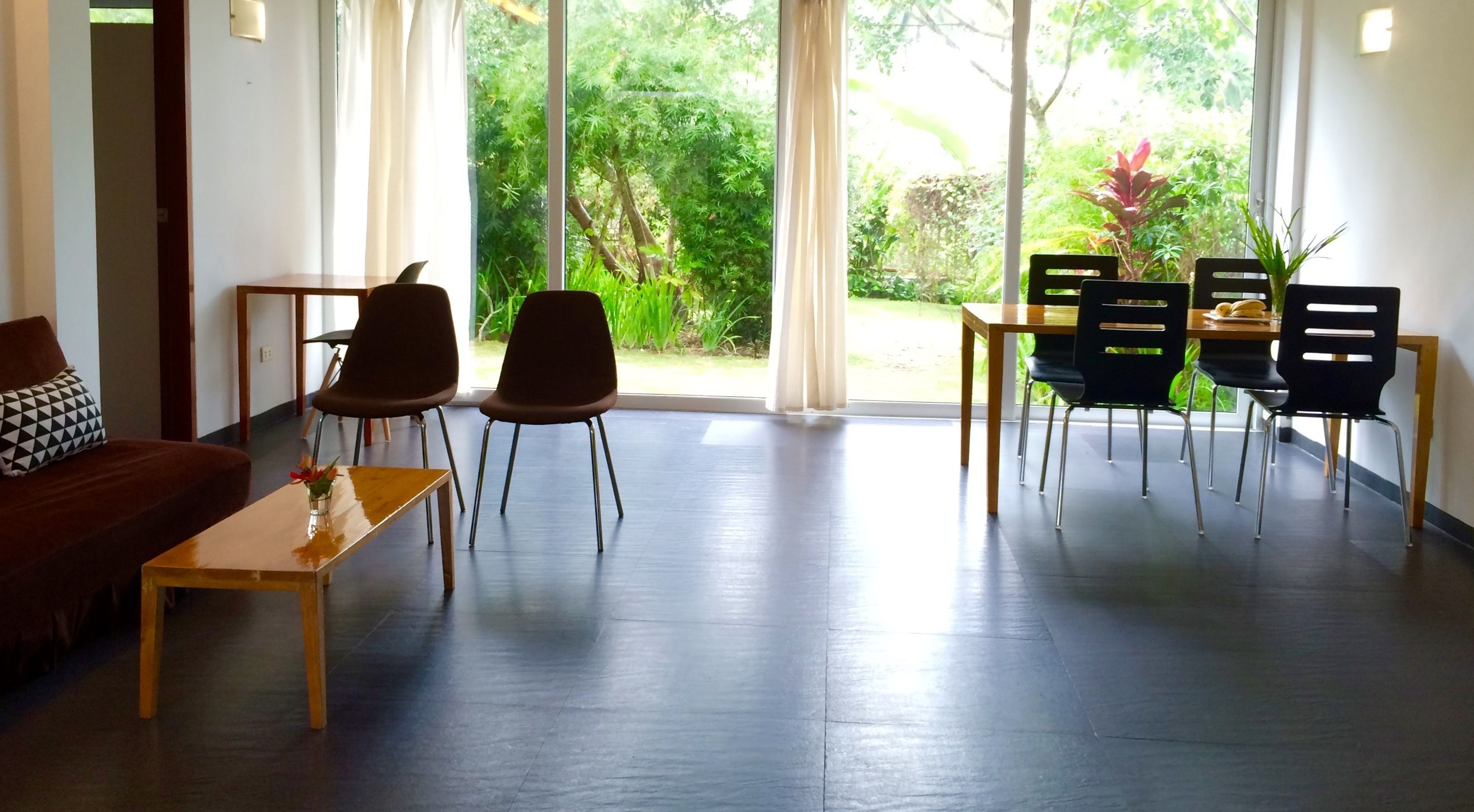 Une chambre d’hôtel moderne avec une table basse en bois, deux fauteuils marron, une table à manger noire avec quatre chaises, et de grandes fenêtres donnant sur un paysage verdoyant.