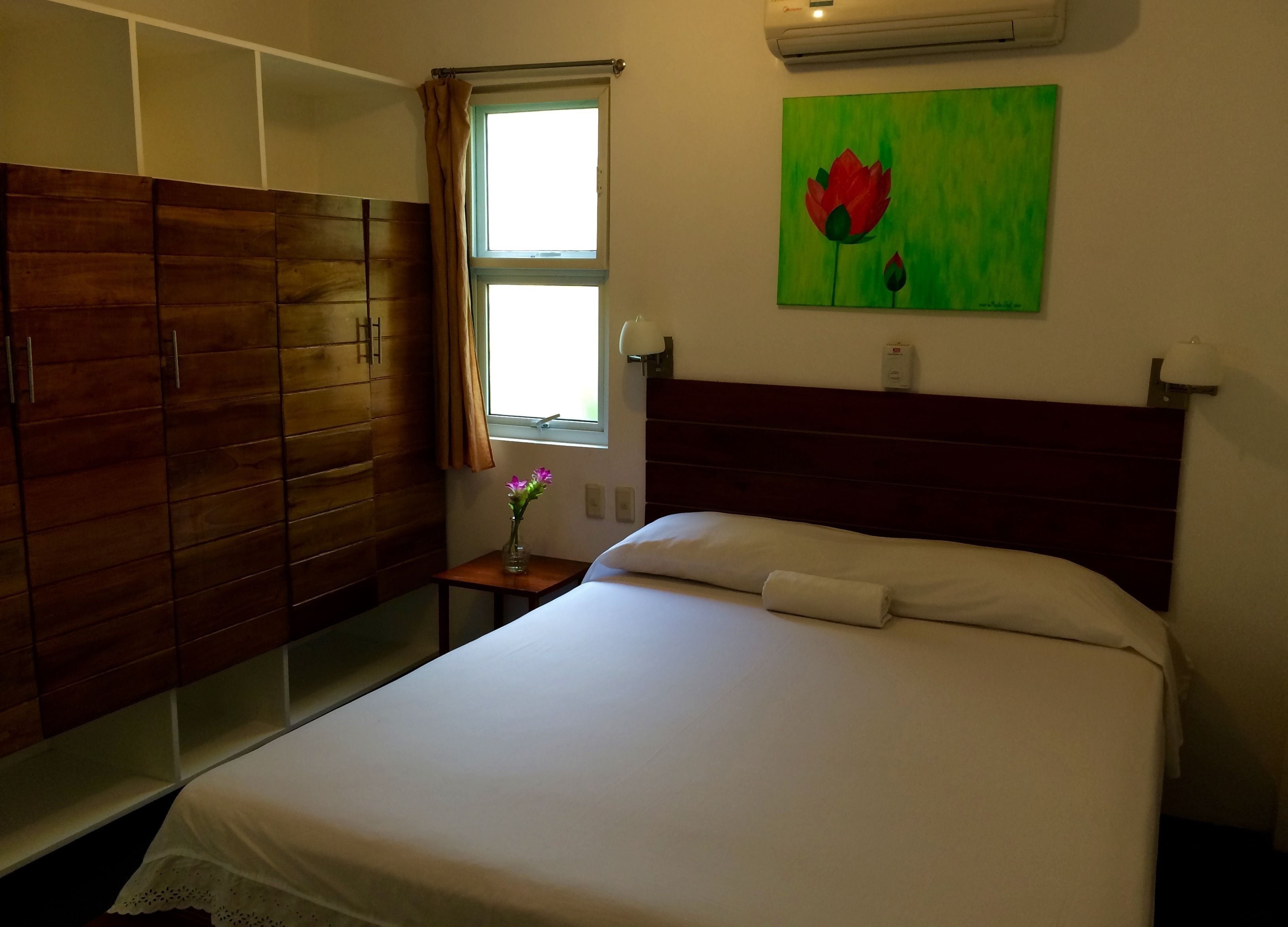 Een hotelkamer met een houten garderobe, een bed met witte lakens, een tafeltje met een bloevas en een schilderij van een bloem aan de muur.