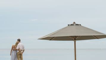 A beach scene with a couple standing under a large umbrella, two wicker lounge chairs, and a calm sea in the background.