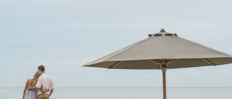 A beach scene with a couple standing under a large umbrella, two wicker lounge chairs, and a calm sea in the background.