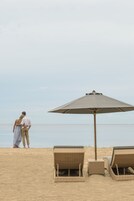 A beach scene with a couple standing under a large umbrella, two wicker lounge chairs, and a calm sea in the background.