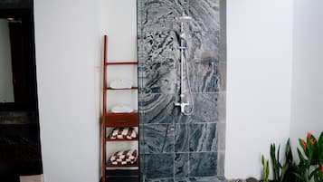 A bathroom with a shower, marble tiles, a wooden shelf with towels, and a small plant area.