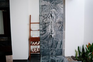 A bathroom with a shower, marble tiles, a wooden shelf with towels, and a small plant area.