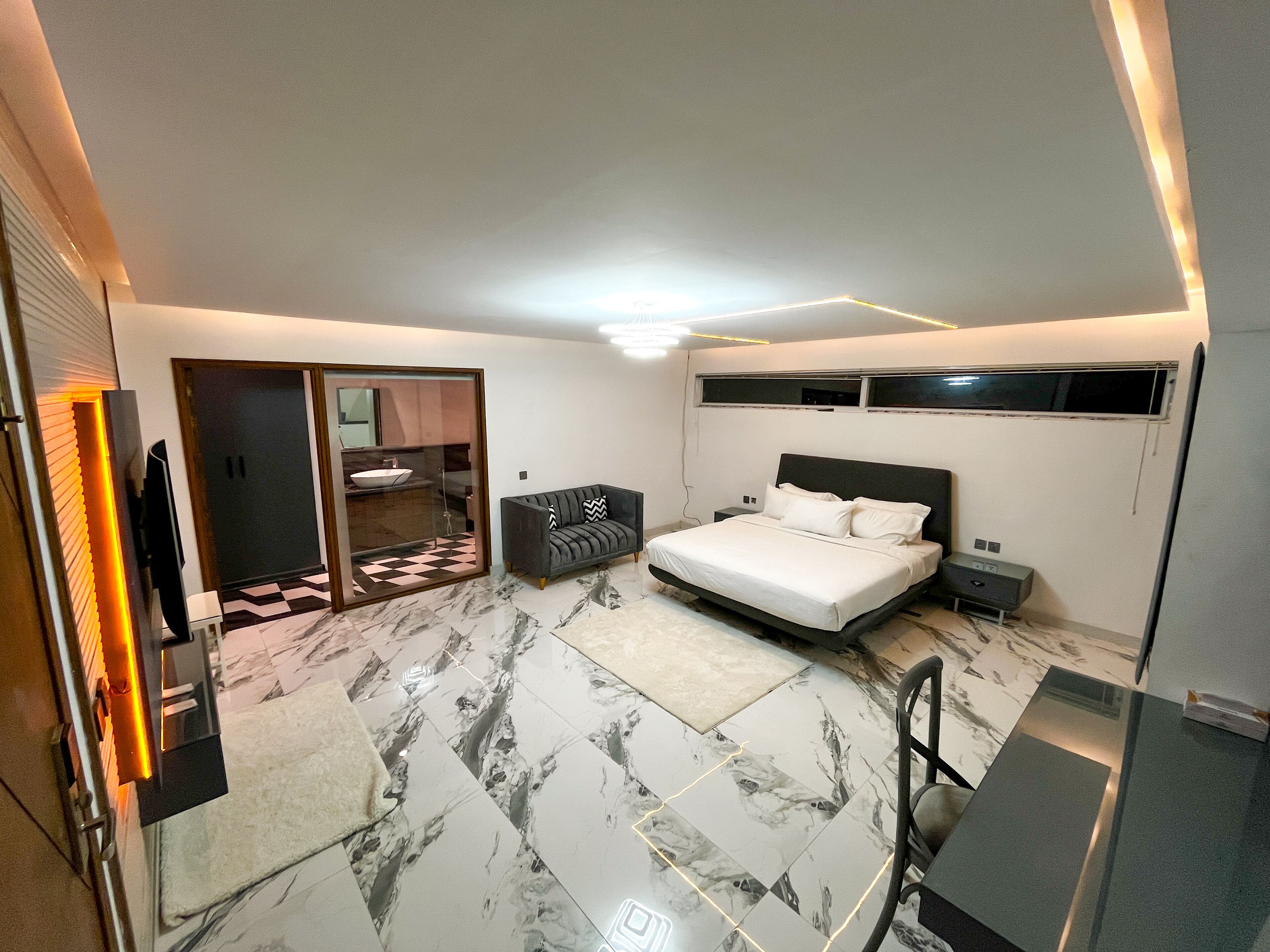 A modern bedroom with a bed, bedside tables, a sofa, a TV, and a bathroom with a sink and mirror.