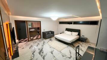 A modern bedroom with a bed, bedside tables, a sofa, a TV, and a bathroom with a sink and mirror.