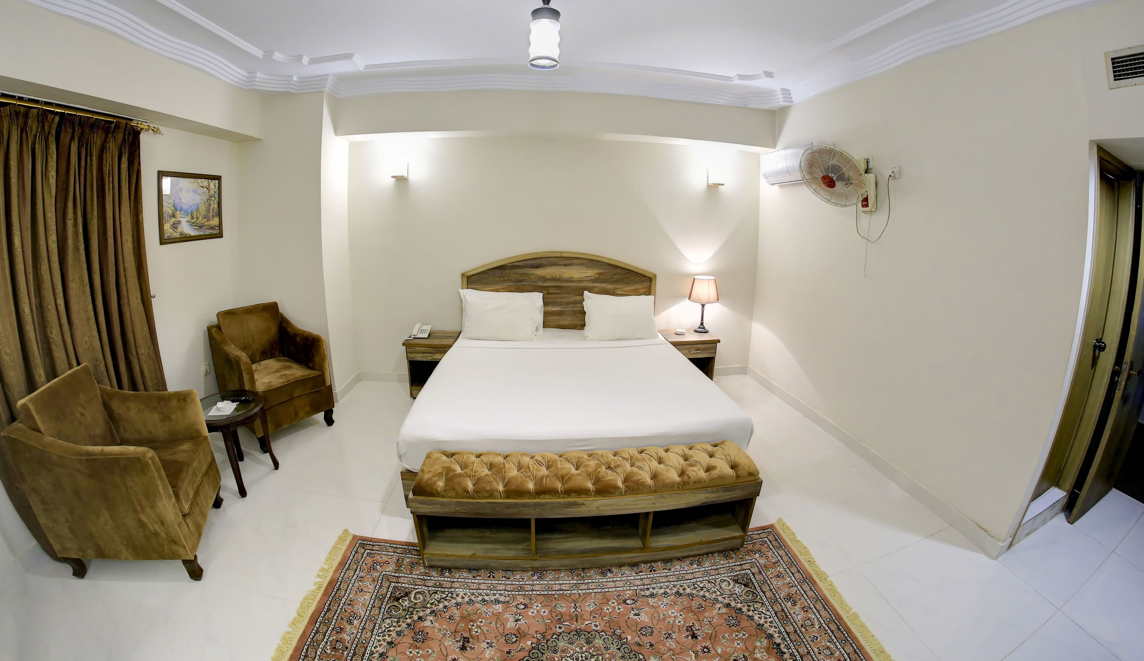 A hotel room with a large bed, a wooden headboard, a bedside table with a lamp, a small nightstand, a brown armchair, a patterned rug, and a painting on the wall.