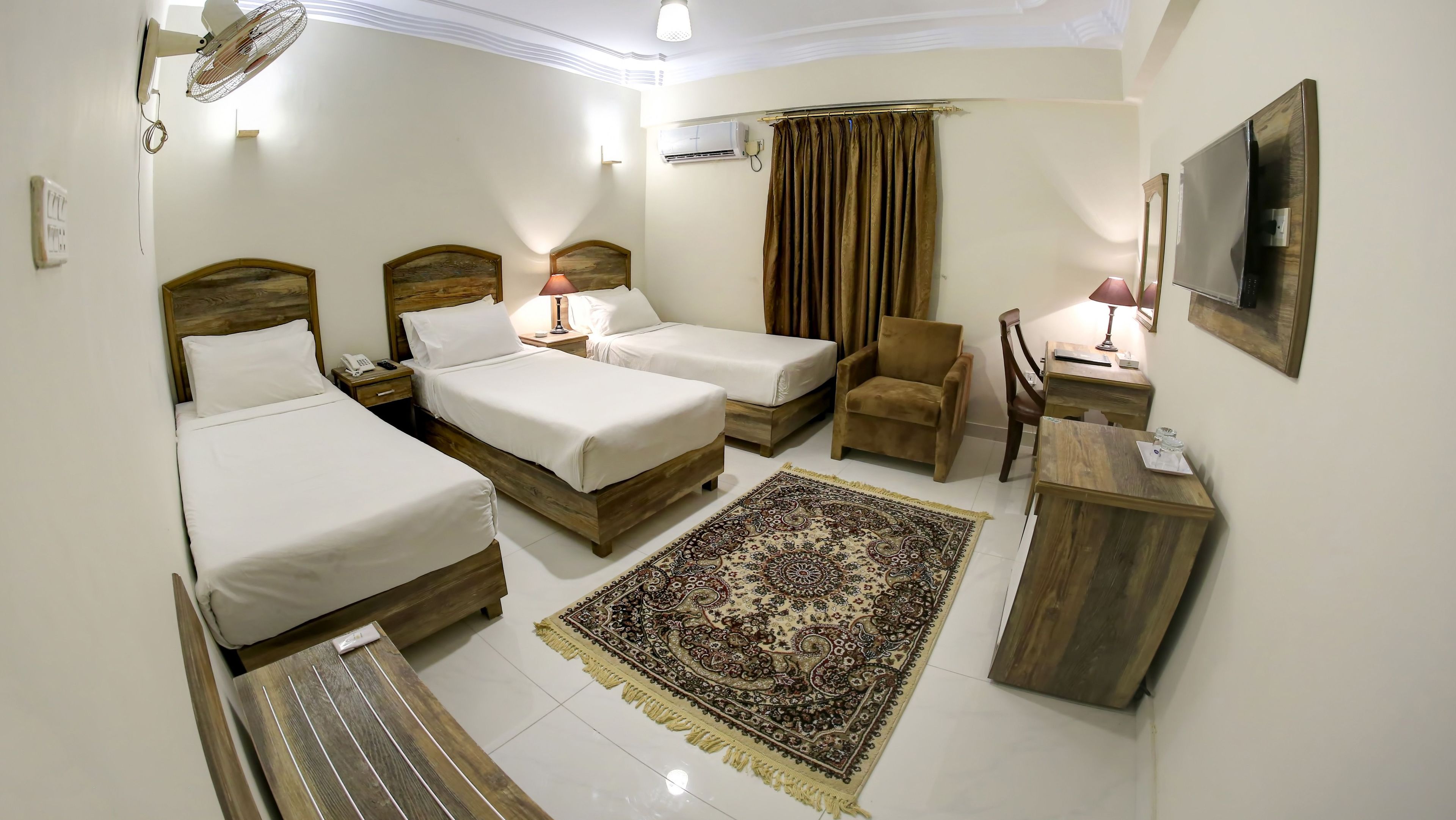 Quarto de hotel com duas camas de solteiro, um sofá, uma cadeira, uma escrivaninha, um tapete e uma TV fixada na parede.