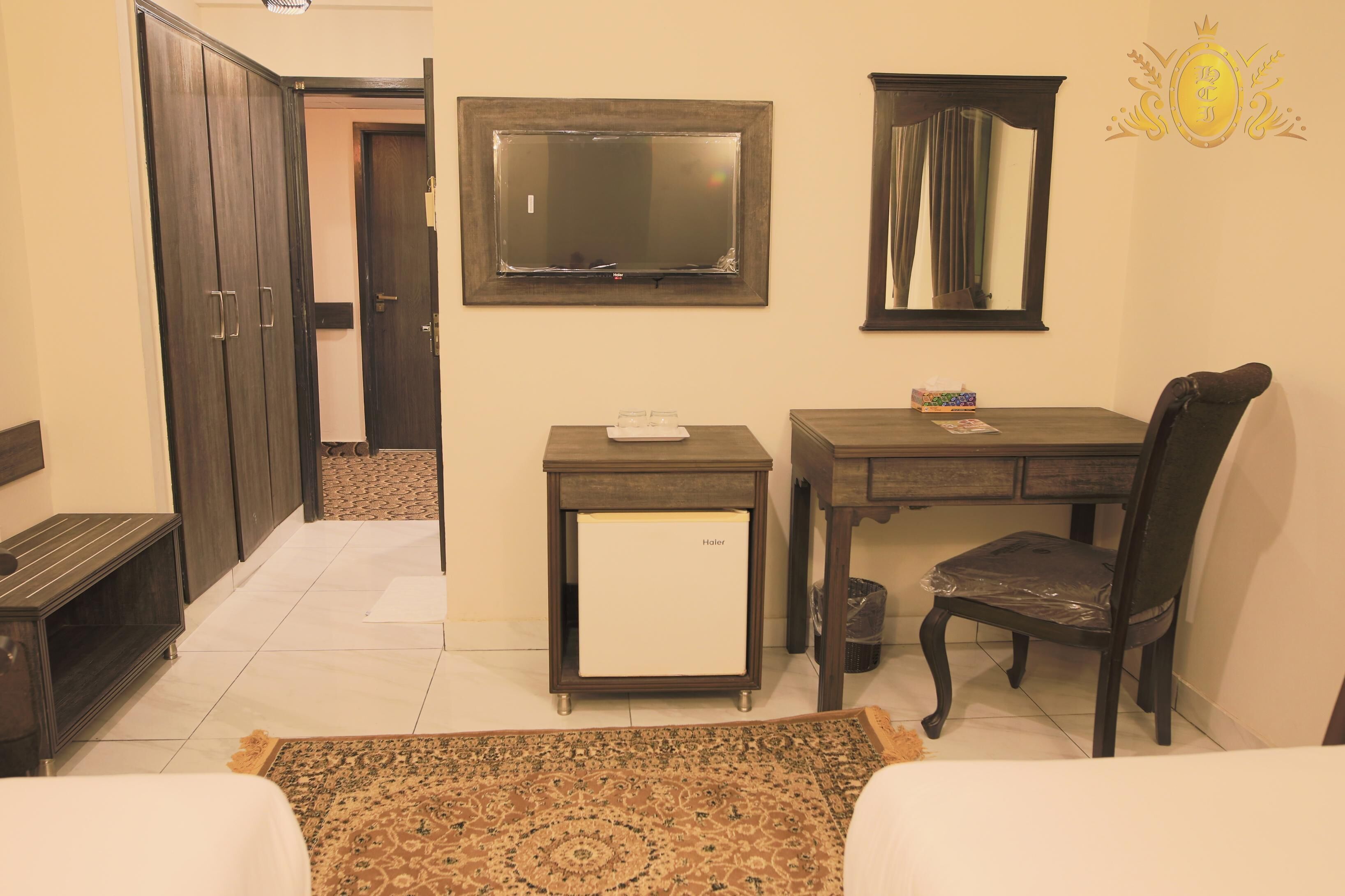 Habitación de hotel con cama, escritorio, silla, refrigerador pequeño, espejo y armario.