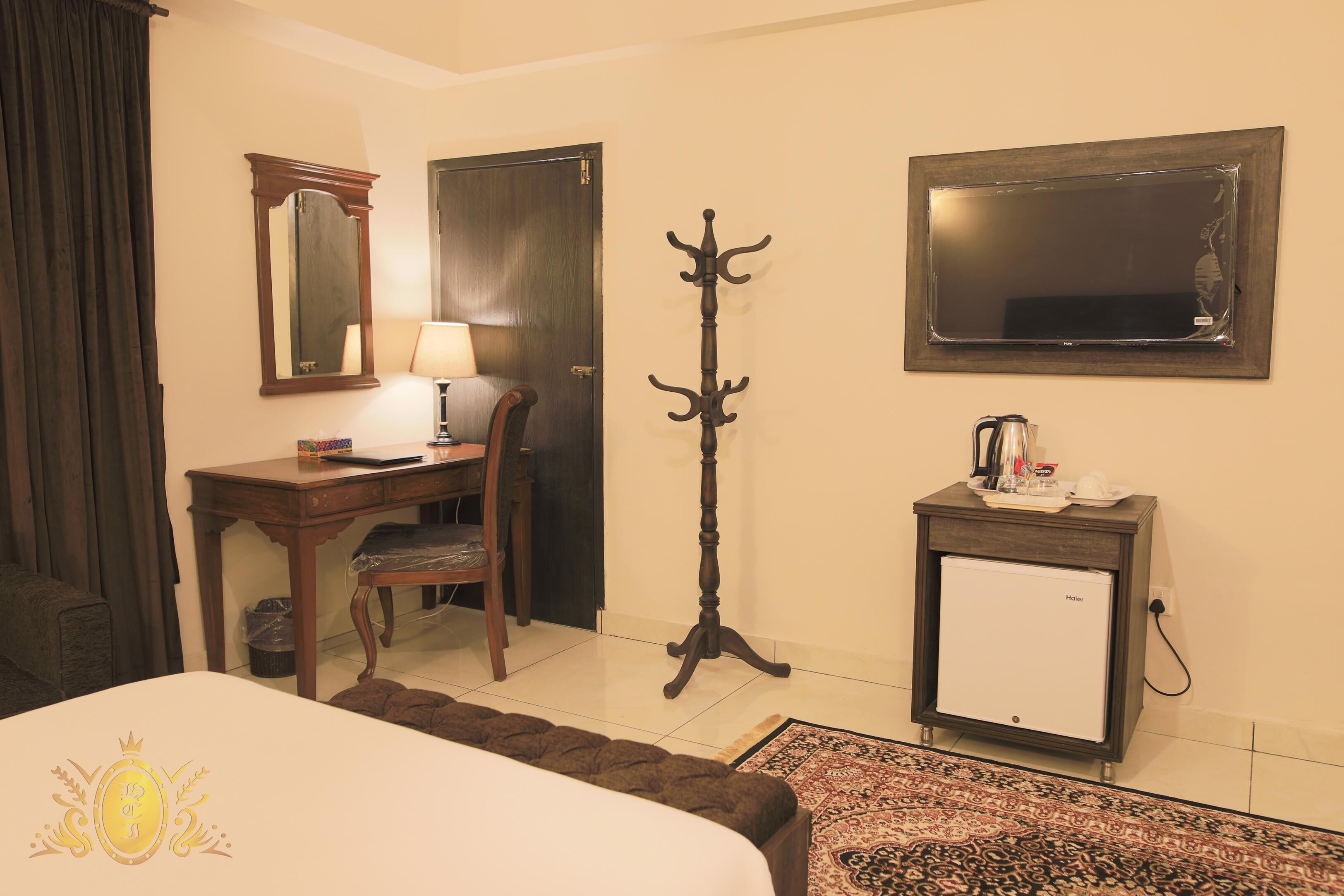 A hotel room with a bed, a desk with a chair, a coat stand, a small refrigerator, and a mirror on the wall.