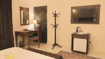 A hotel room with a bed, a desk with a chair, a coat stand, a small refrigerator, and a mirror on the wall.