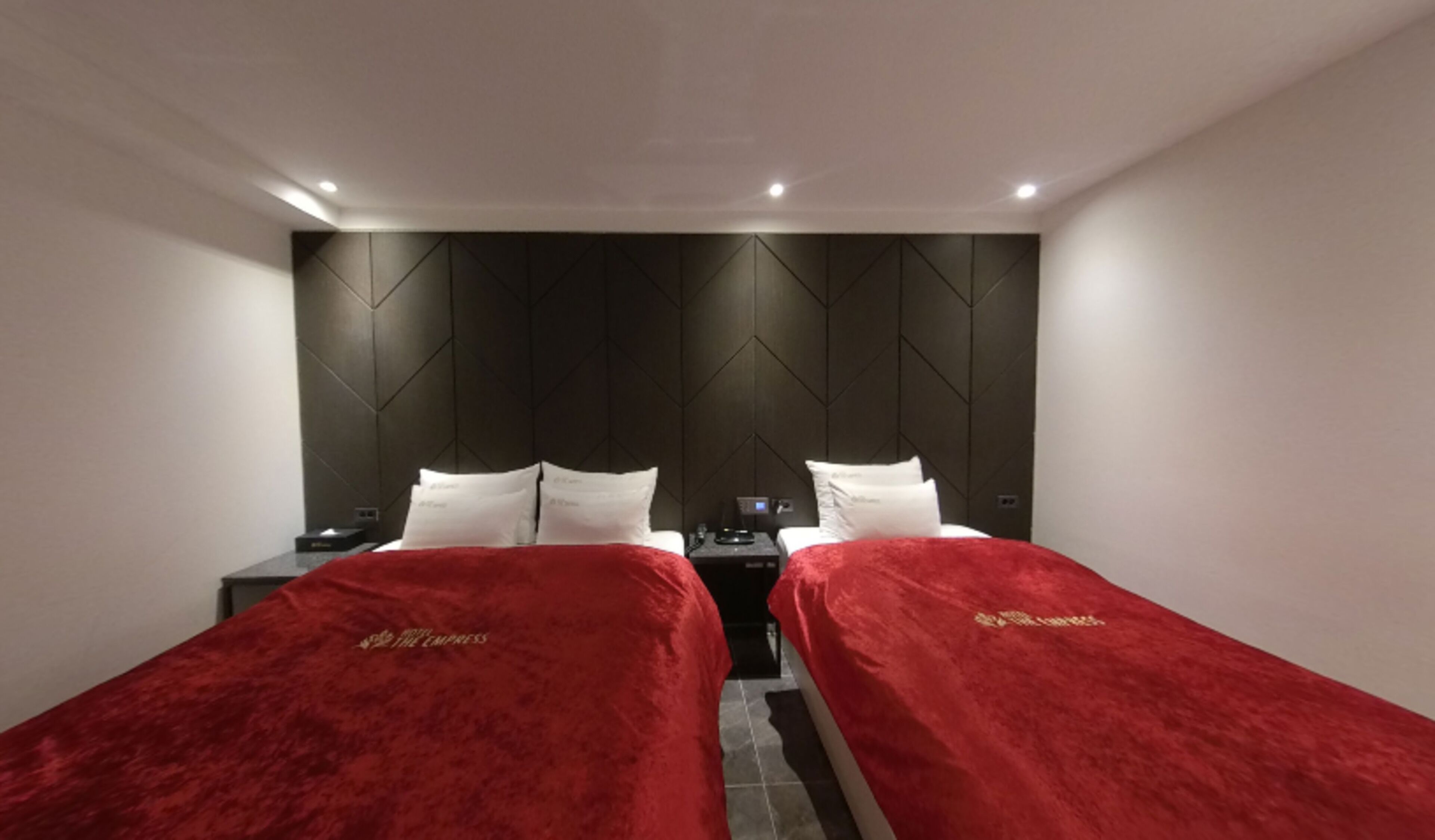 Habitació d'hotel amb dos llits, cadascun amb llençol i almoines vermelles, un capçal fosc i una petita taula de nit amb telèfon.