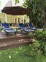 A patio with three lounge chairs under a large umbrella, surrounded by lush greenery and a building with a tiled roof.