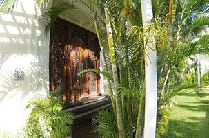 A traditional wooden door with intricate carvings, surrounded by lush greenery and a white wall.