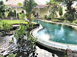 A pool with a curved edge, surrounded by various plants and a building with a tiled roof in the background.