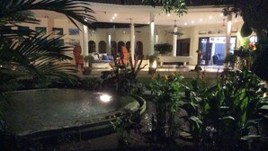 A courtyard with a central water feature, surrounded by potted plants and a building with arched windows. - Villa Costa Sanur (Denpasar)