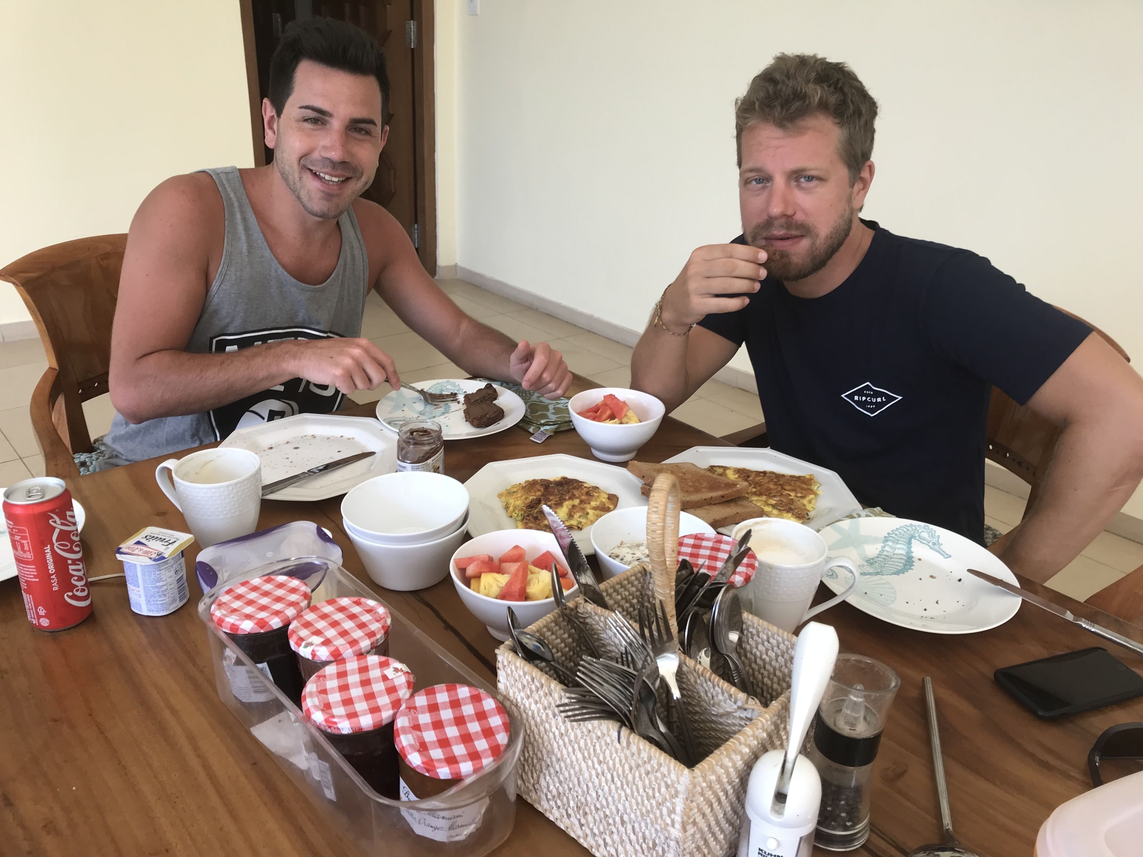 Two people sitting at a wooden table with various food items, utensils, and condiments.