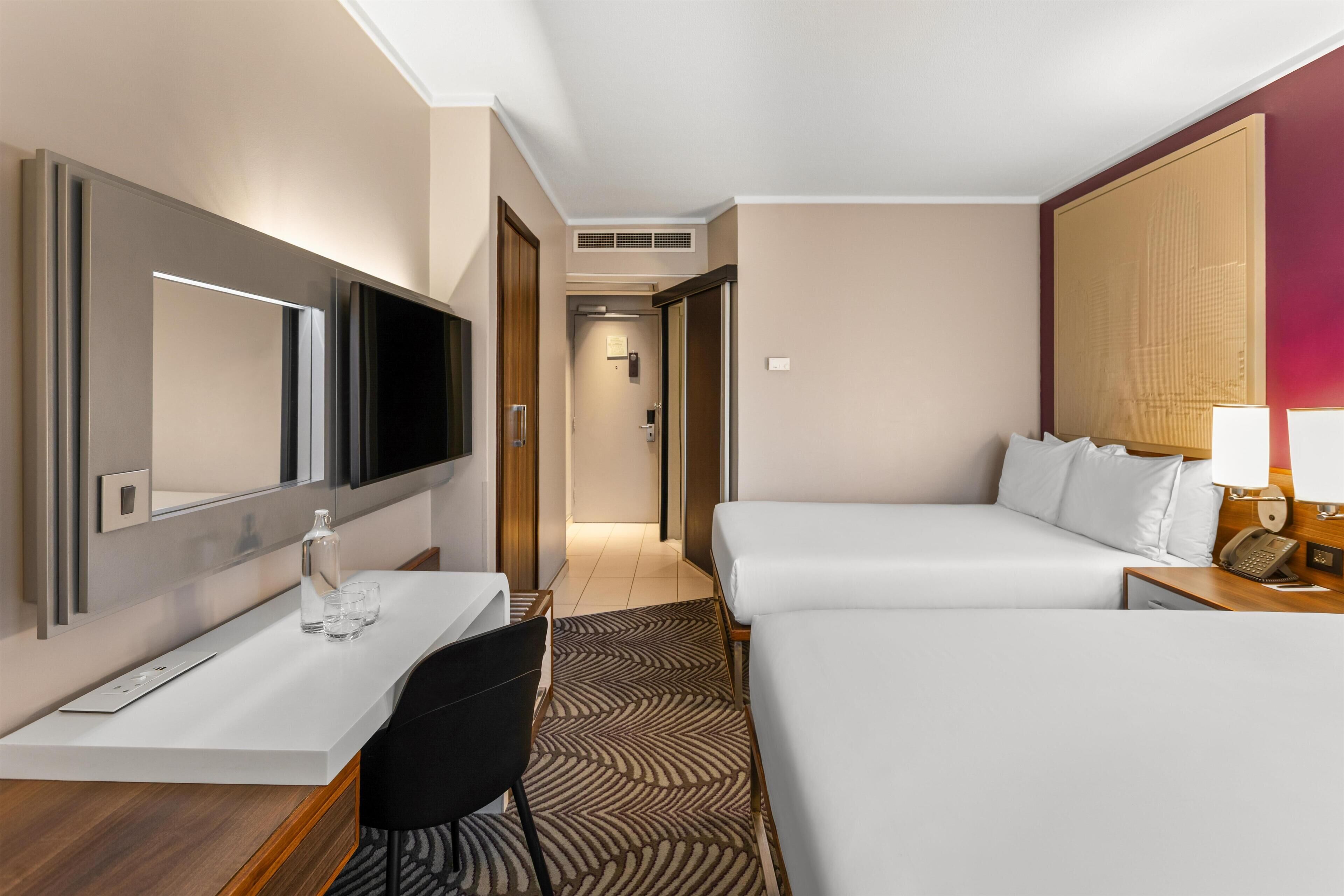 A hotel room with a bed, a desk, a chair, a TV, a mirror, and a nightstand with a lamp.