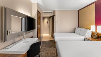 Habitación de hotel con cama, escritorio, silla, televisor, espejo y mesita de noche con lámpara.