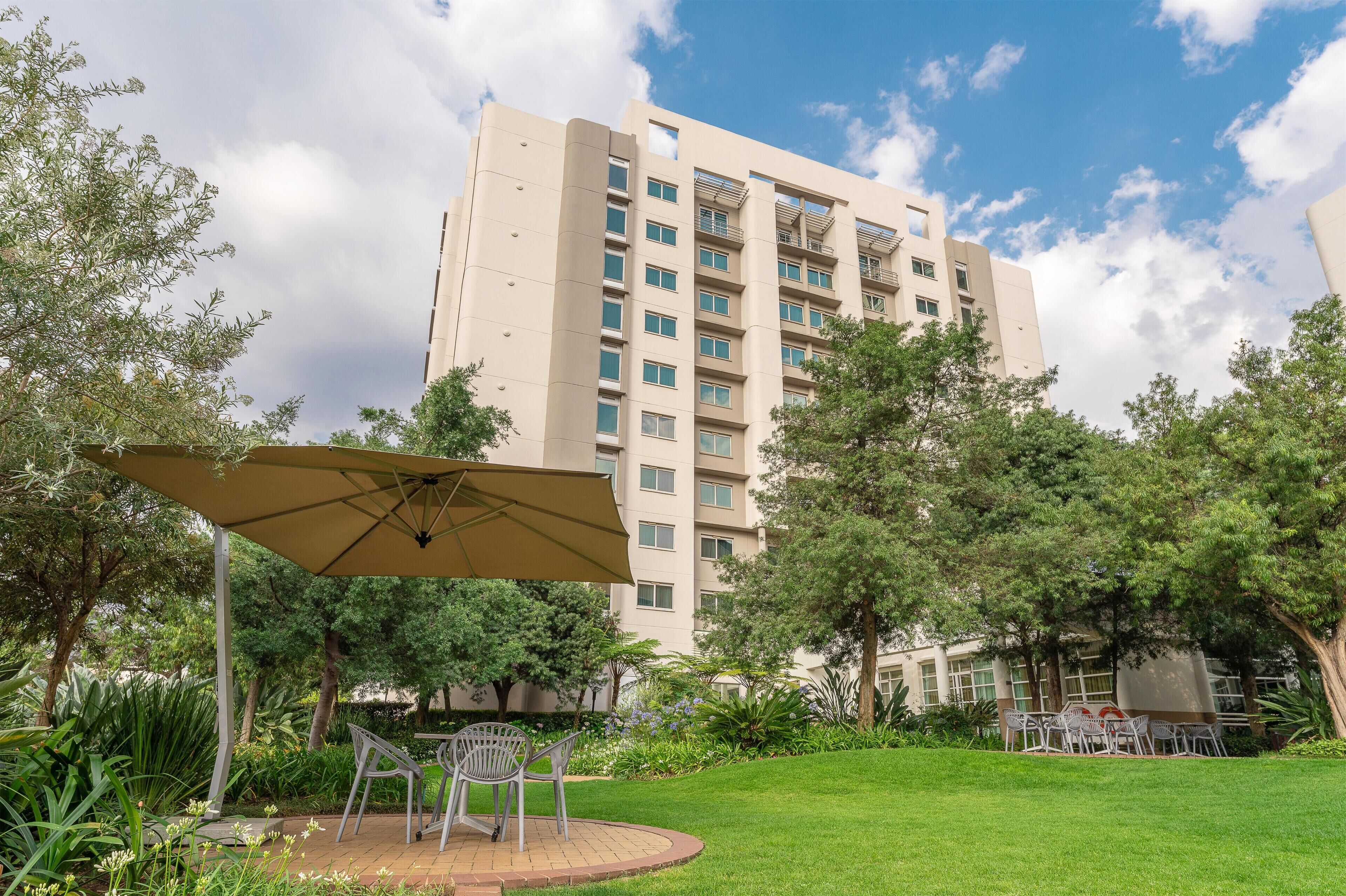 Un patio con zona de comedor, rodeado de vegetación y un edificio alto al fondo.
