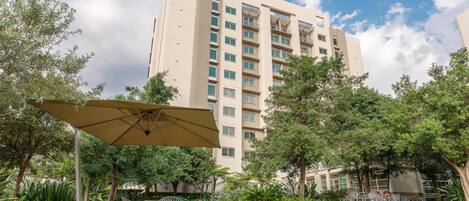 A courtyard with a dining area, surrounded by greenery and a tall building in the background.