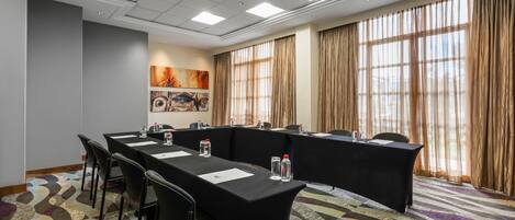 Uma sala de conferências com uma mesa comprida, cadeiras e uma grande janela com cortinas.