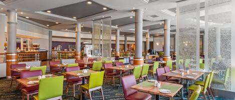 A spacious hotel dining area with multiple tables and chairs, a bar, and a kitchen in the background.