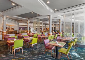 A spacious hotel dining area with multiple tables and chairs, a bar, and a kitchen in the background.