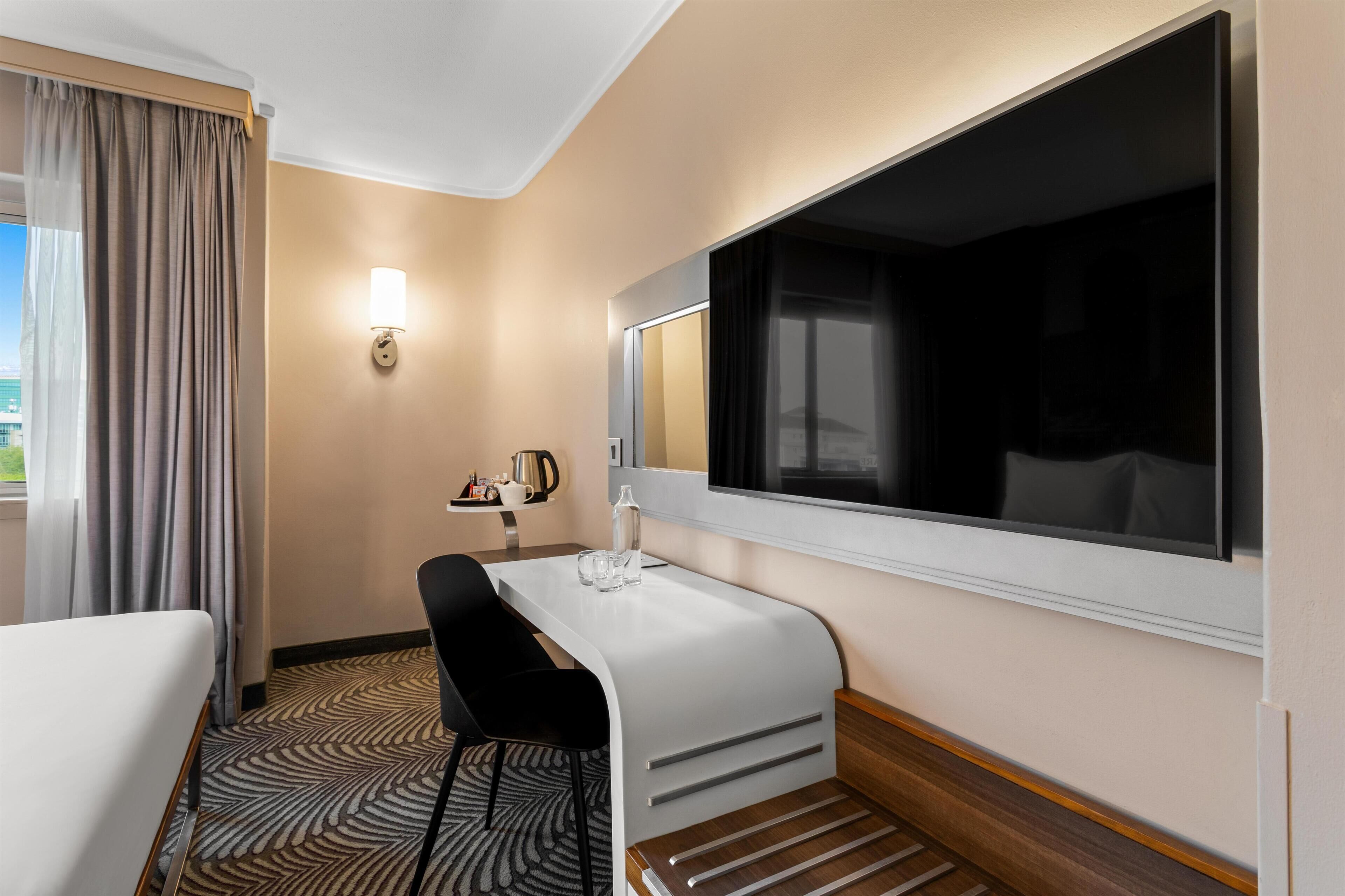 A hotel room with a bed, a desk with a chair, a large window, and a wall-mounted TV.