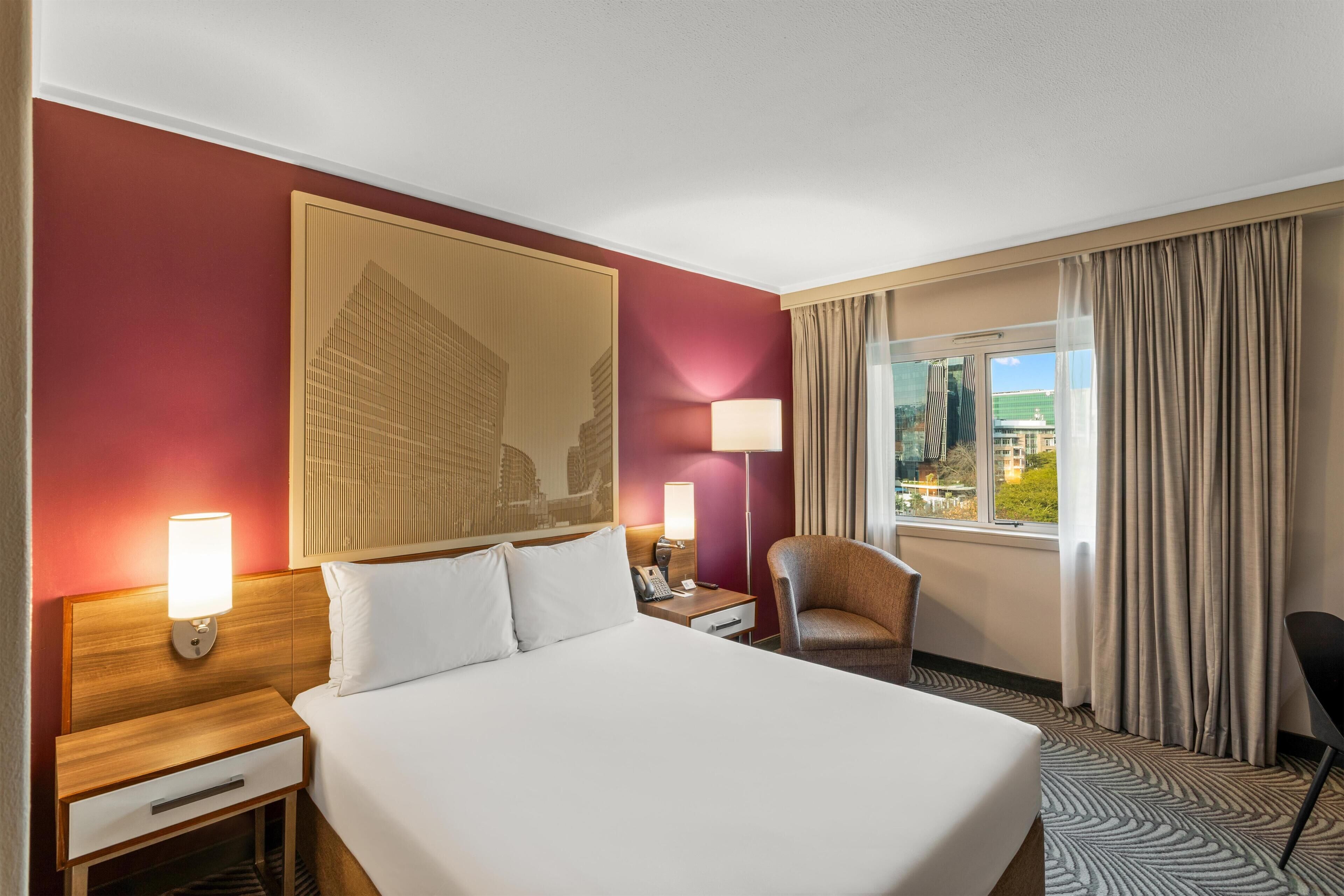 Una camera d'albergo con un letto grande, un comodino con una lampada, una sedia, una scrivania con un telefono e una finestra con tende.
