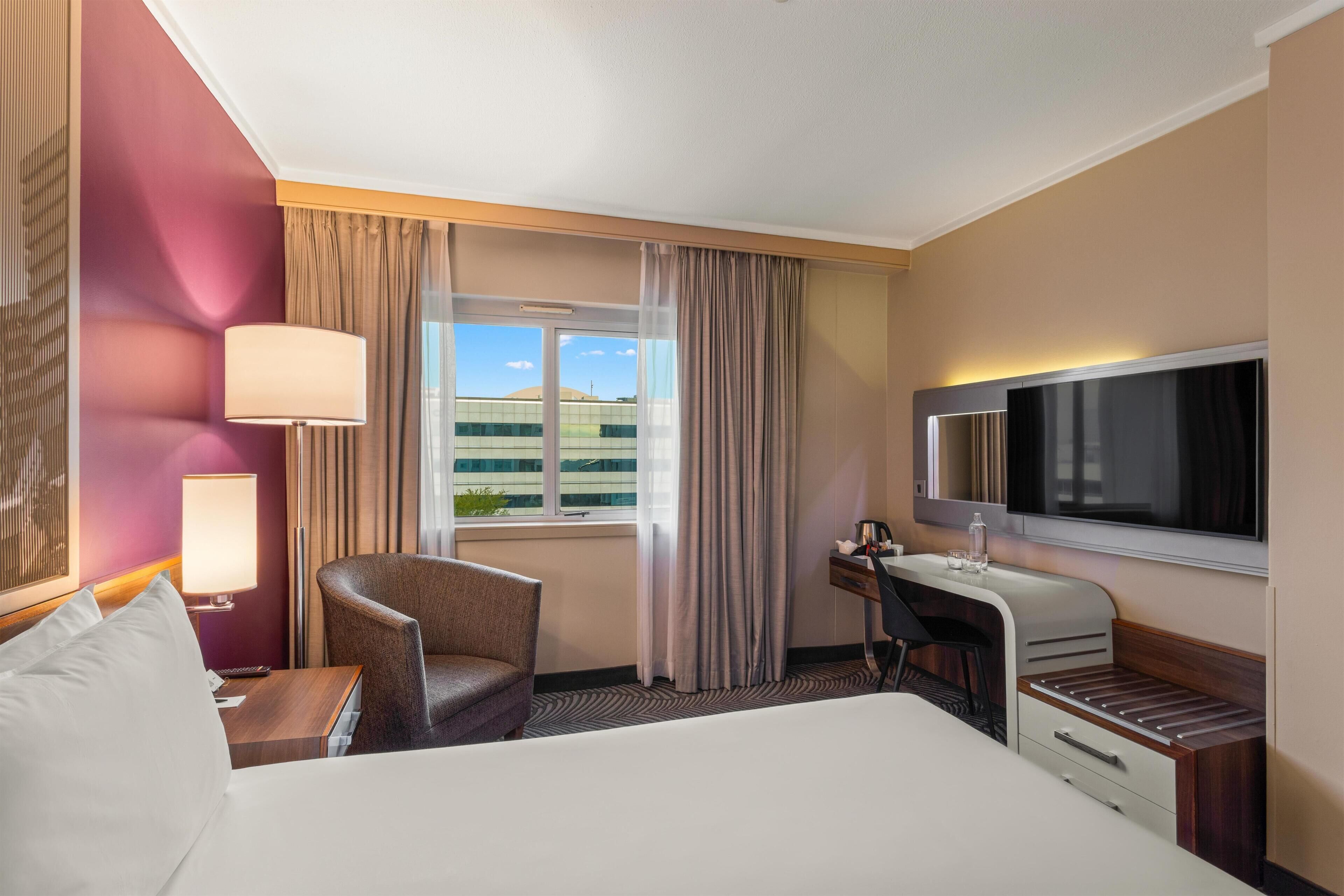 Habitación de hotel con cama, escritorio, silla, televisor y ventana con cortinas.
