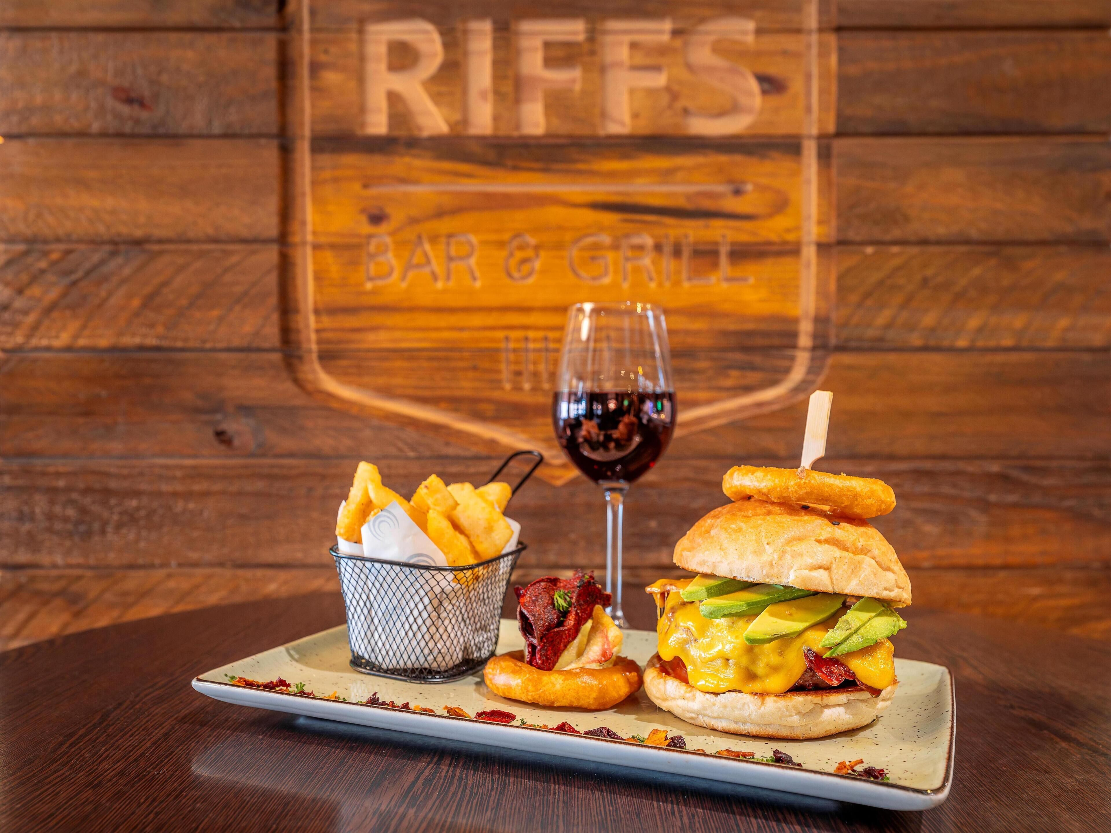 A meal with a burger and fries on a plate, a glass of red wine, and a menu board in the background.
