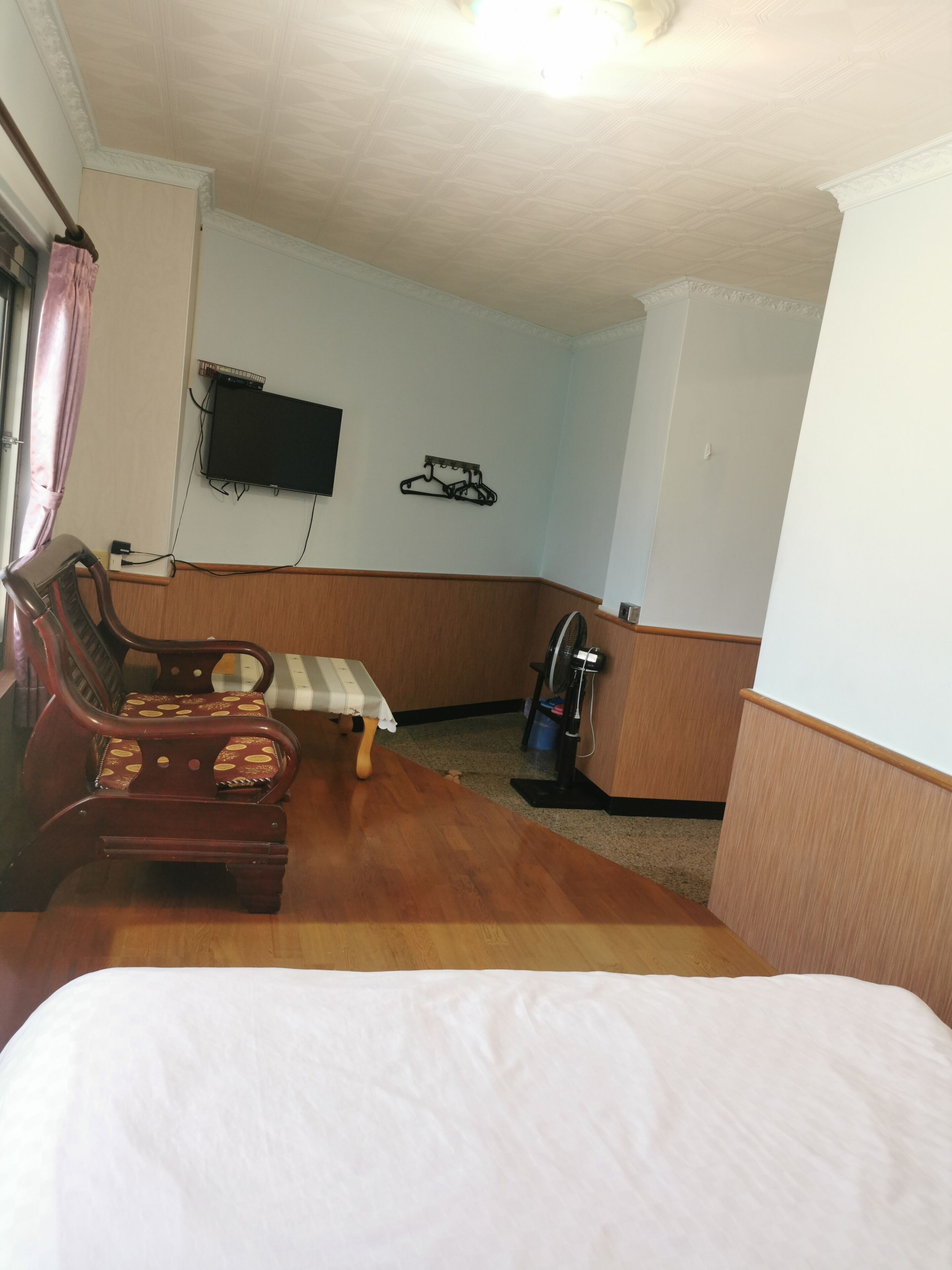 A hotel room with a bed, a wooden chair with a patterned cushion, a small table, a wall-mounted TV, and a wall-mounted drying rack.