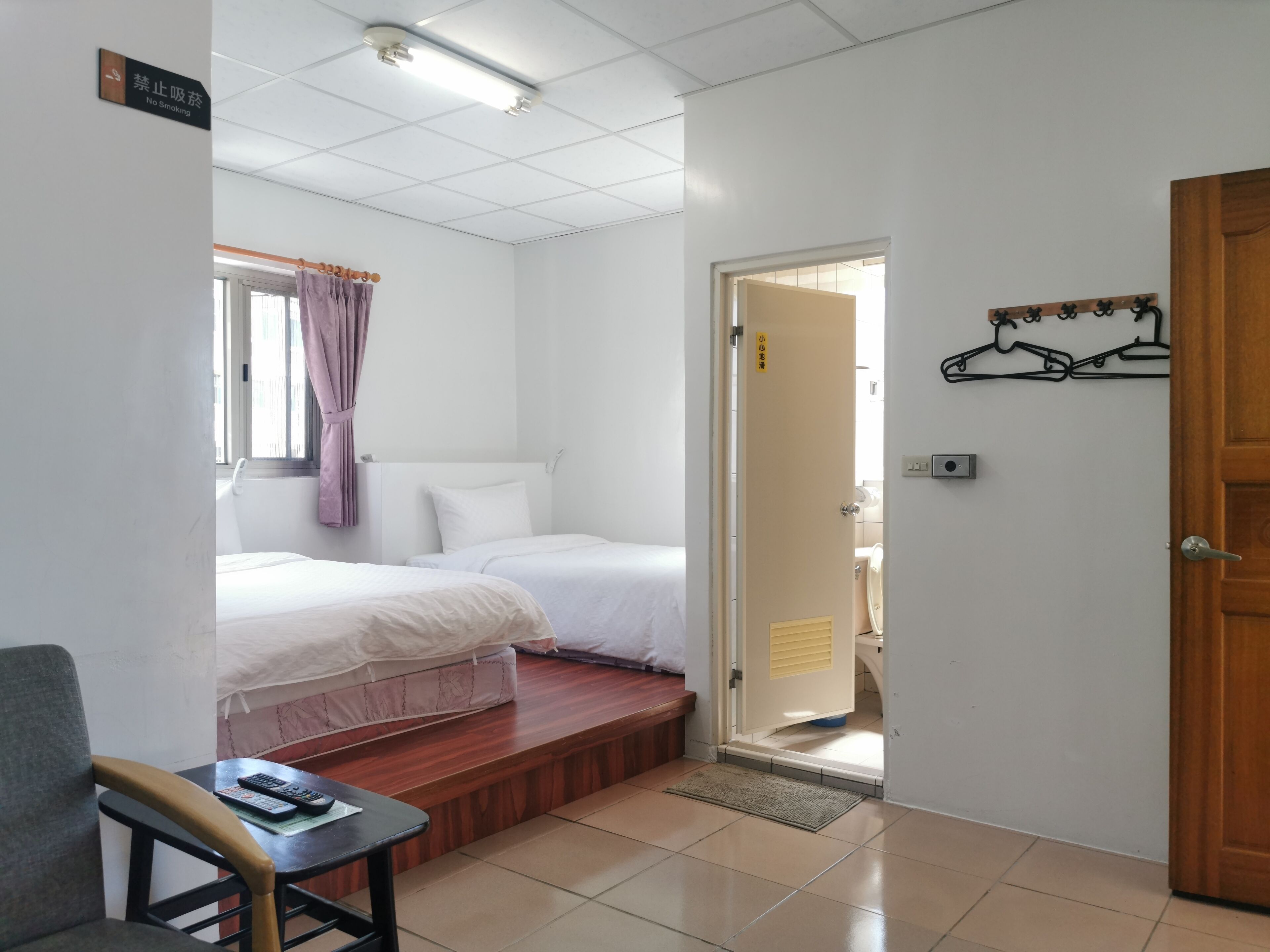 Habitación de hotel con dos camas, una mesita con un control remoto, un banco de madera y una puerta que da al baño.