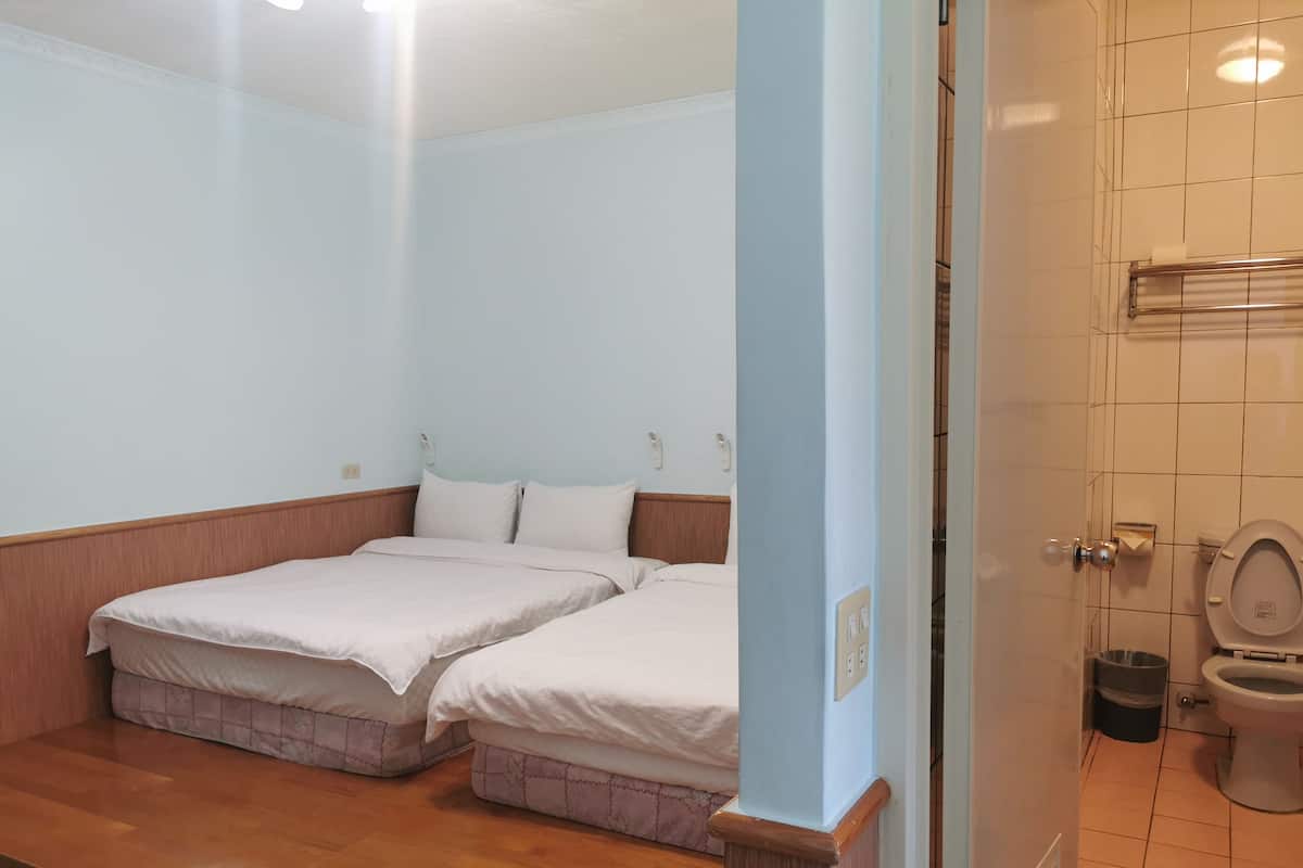 A small, clean room with two beds, a wooden floor, a light blue wall, and a bathroom with a toilet and sink.
