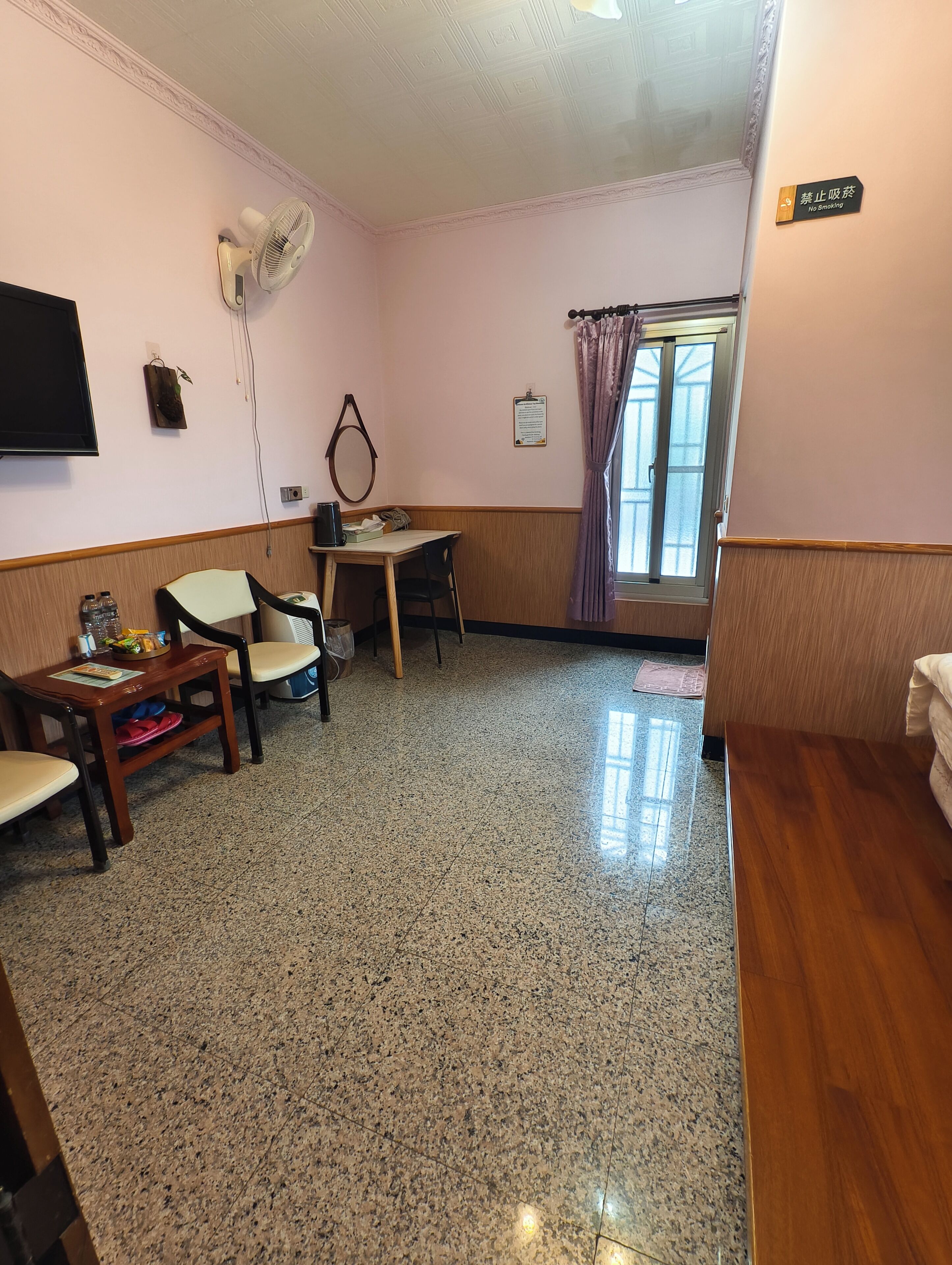 Une chambre d’hôtel comprenant un coin repas, une télévision, un ventilateur, une fenêtre avec des rideaux et un panneau indiquant « Il est interdit de fumer ».
