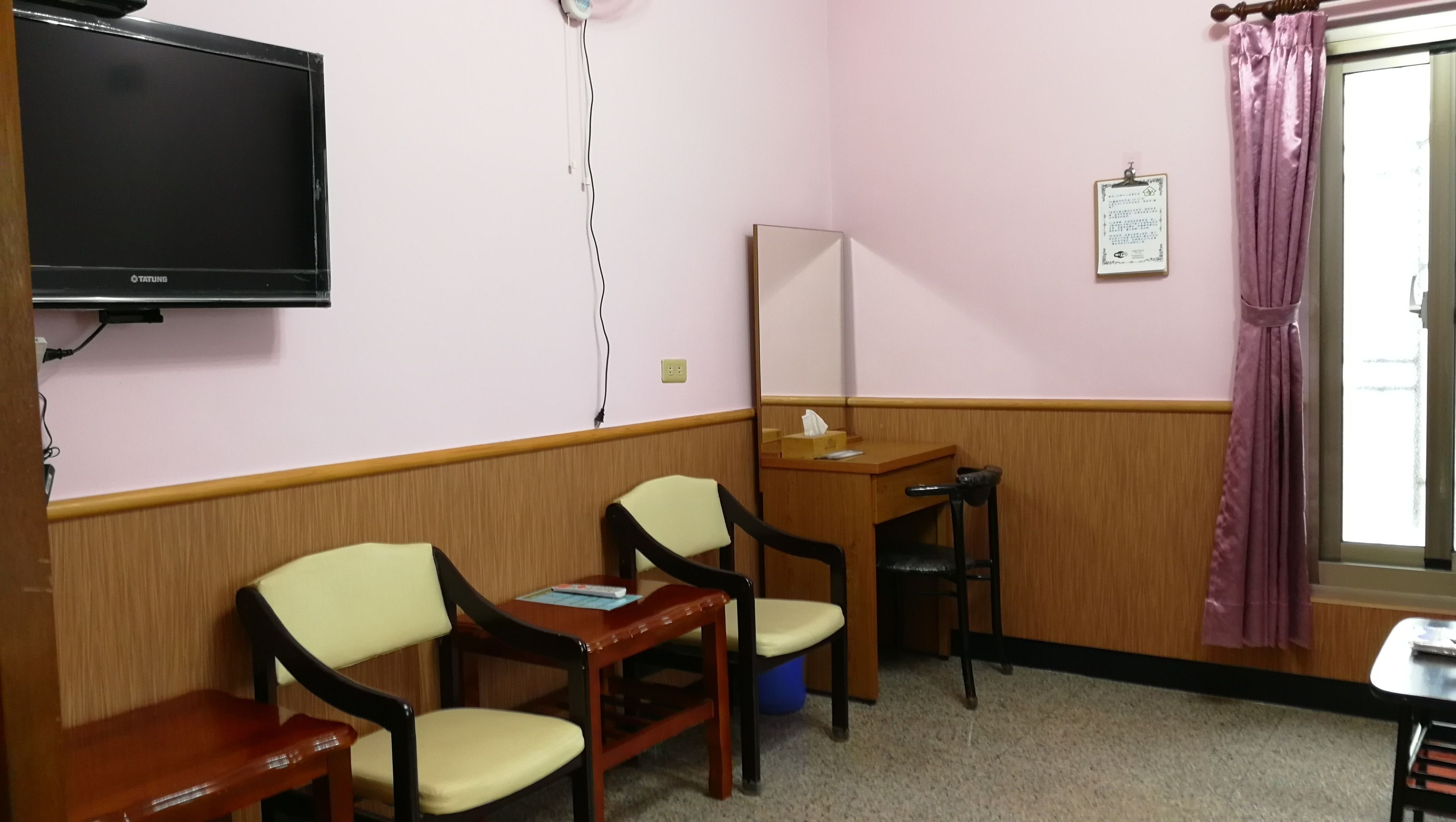 A room with two wooden chairs, a small table, a TV mounted on the wall, a window with a curtain, and a mirror on the wall.