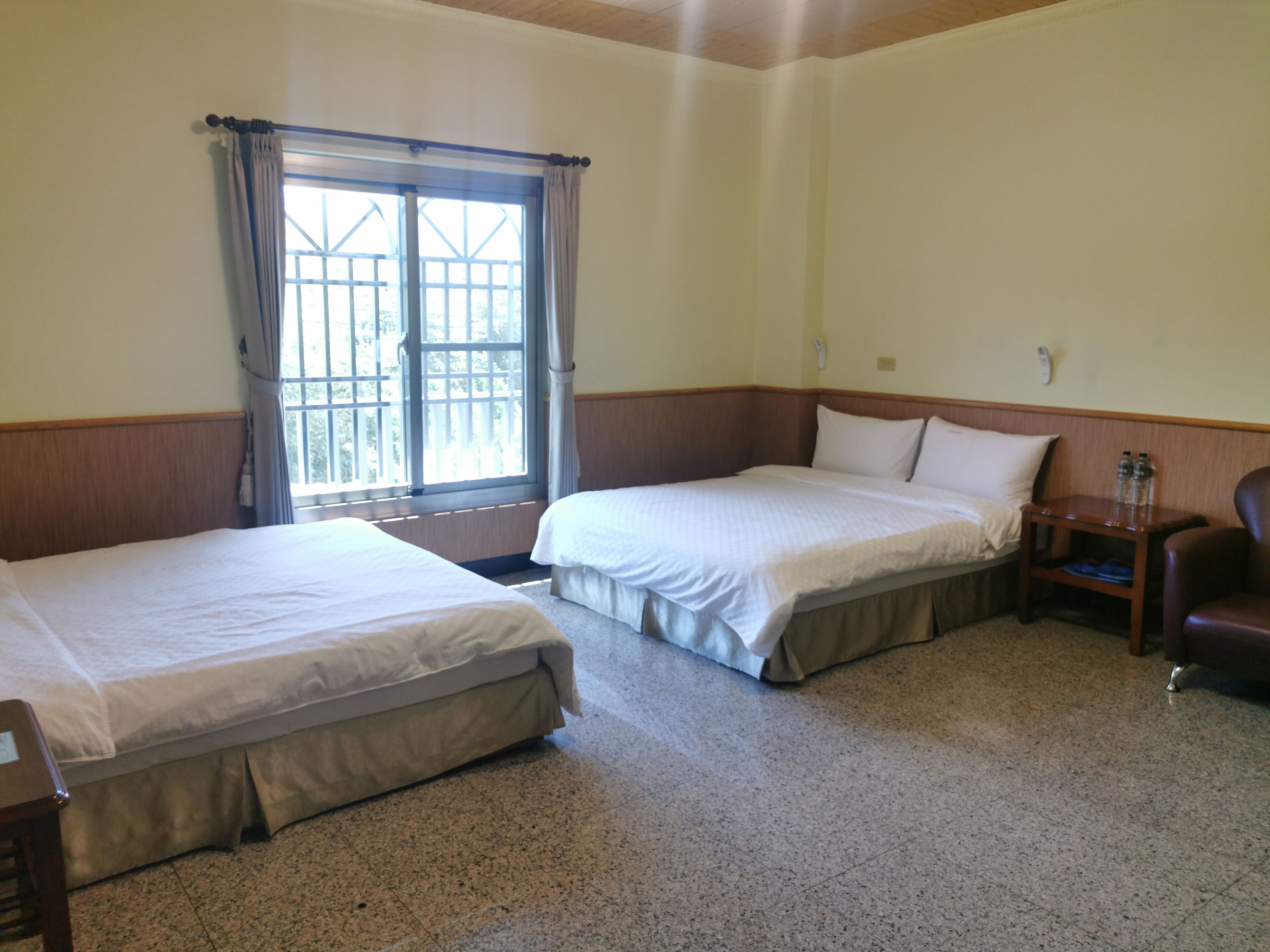 Habitación de hotel con dos camas individuales, cabecero de madera, mesita de noche pequeña con lámpara y ventana con cortinas.