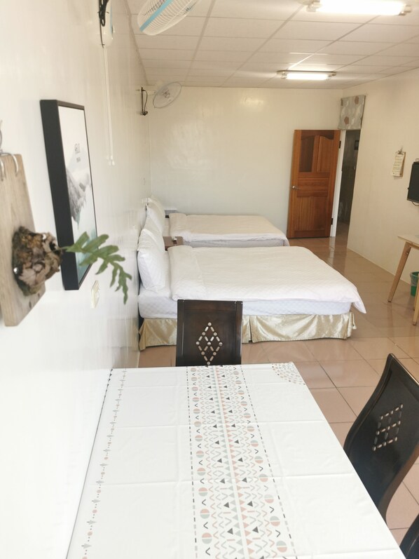 A hotel room with two beds, a dining table, chairs, a wooden door, and a wall-mounted fan.