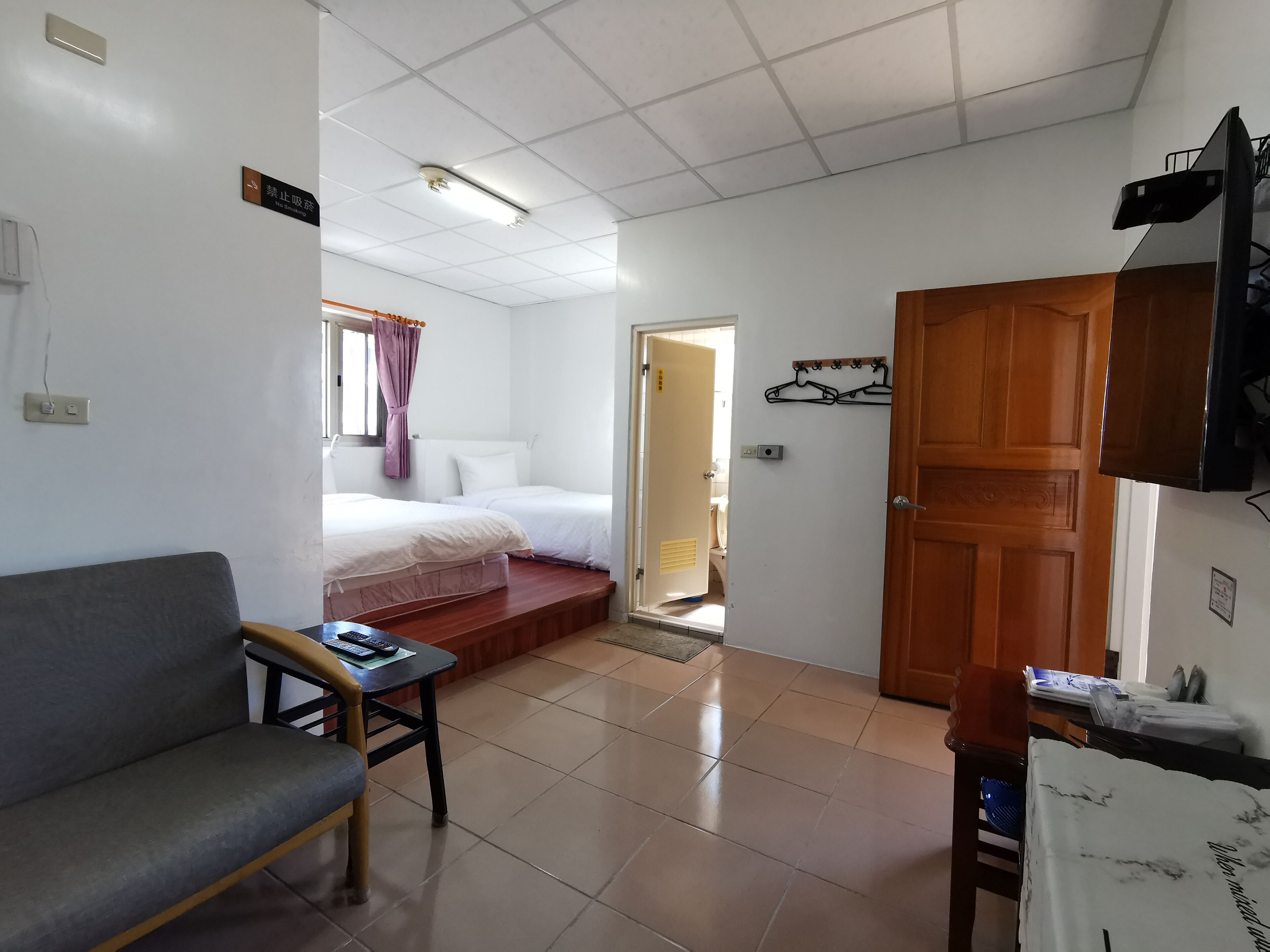 A compact hotel room with a bed, a small table with a chair, a wooden door, and a wall-mounted TV.