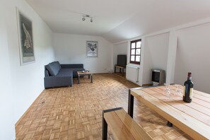 A modern living room with a herringbone wooden floor, a grey sofa, a wooden coffee table, a TV, and a dining table with a bottle and glasses.