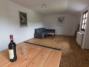 A room with a wooden table, a bottle of wine, and a glass. There is a grey sofa and a small table in the background.