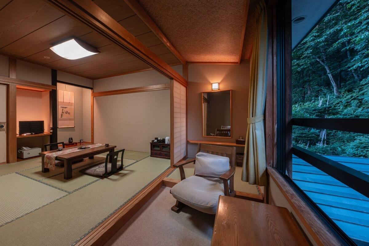 A traditional Japanese room with tatami flooring, a low table, a chair, a television, and a view of a forest through a window.