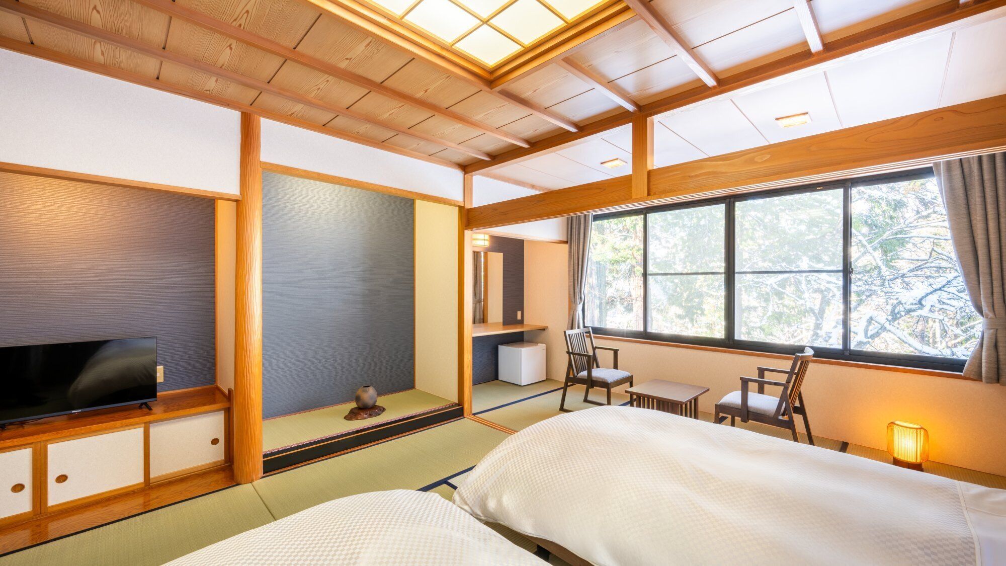 Una camera d'albergo in stile tradizionale giapponese, con pavimento in tatami, televisione, un tavolino e una finestra vista alberi.