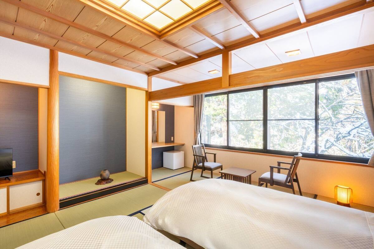 A traditional Japanese-style hotel room with tatami flooring, a television, a small table, and a window with a view of trees.
