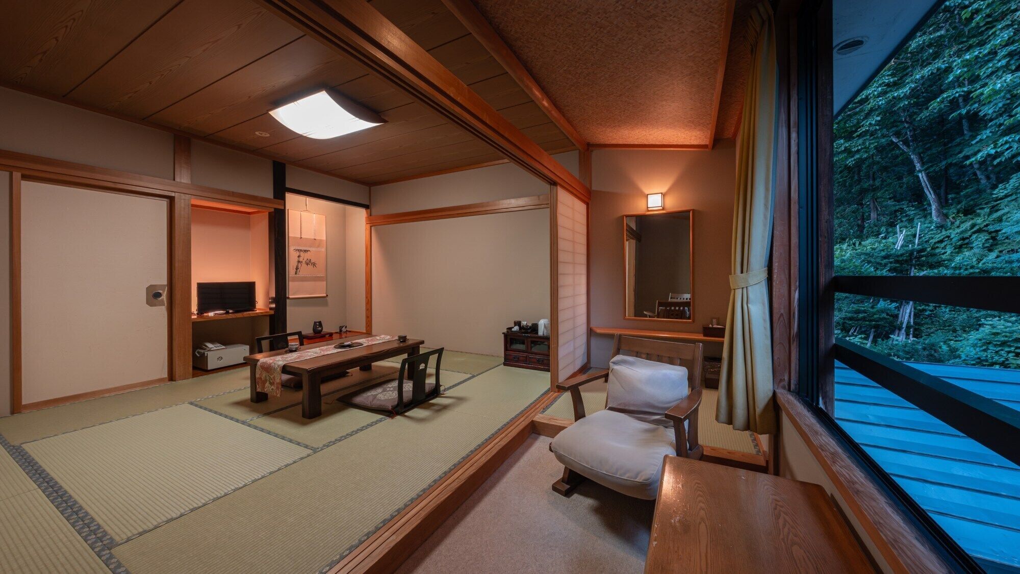 Una stanza tradizionale giapponese con pavimento in tatami, un tavolino, una sedia, un televisore e vista su una foresta attraverso una finestra.