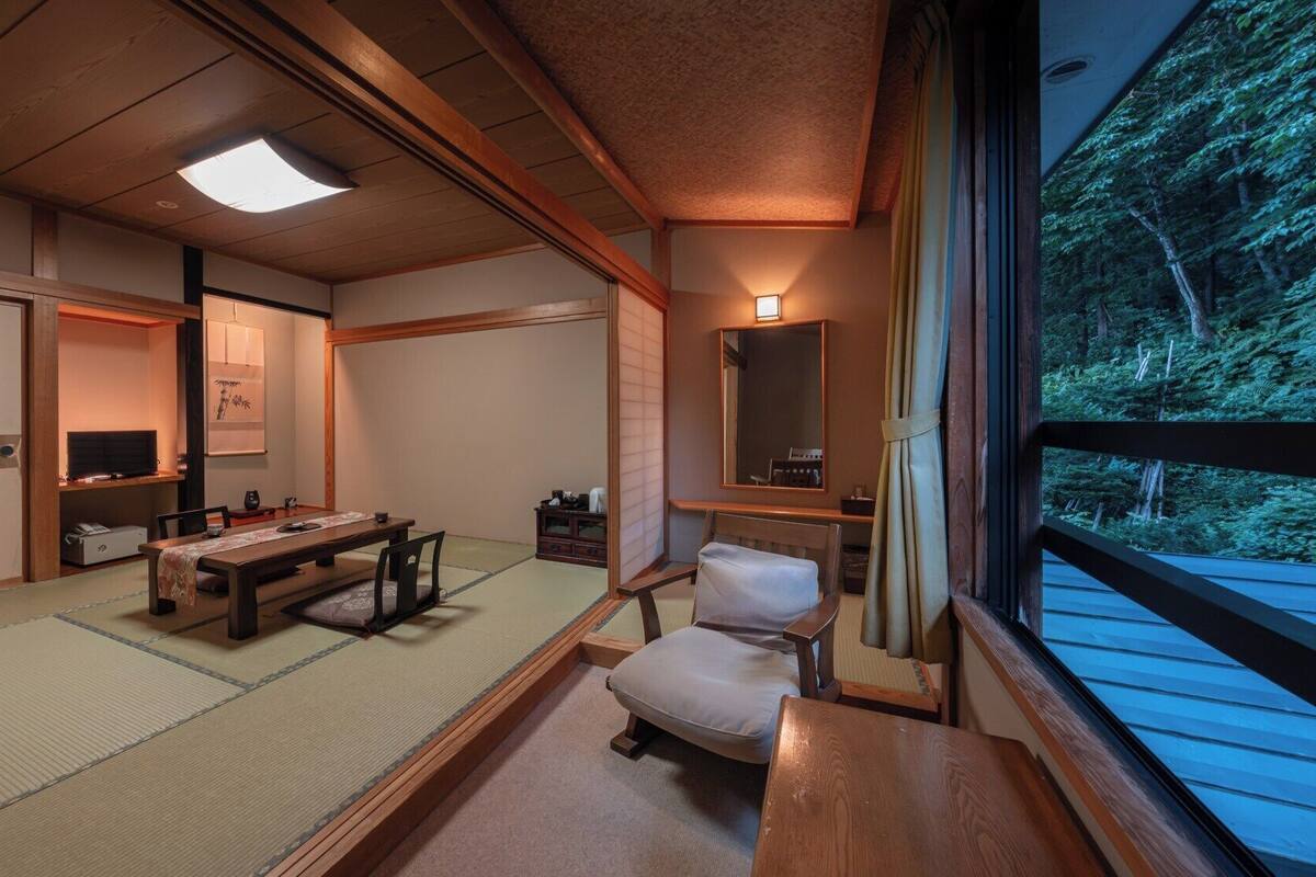 A traditional Japanese room with tatami flooring, a low table, a chair, a television, and a window with a view of trees.